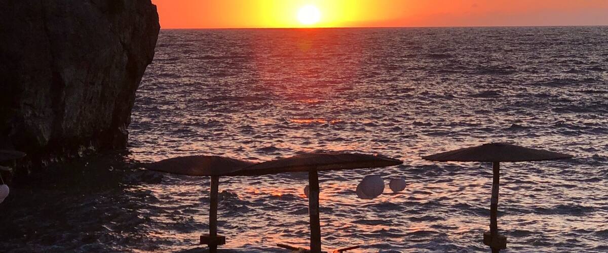 A sunset over the ocean with a rocky cliff and beach umbrellas.