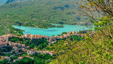 A mountainous village with terracotta-roofed buildings nestled among lush green hills and a turquoise lake.