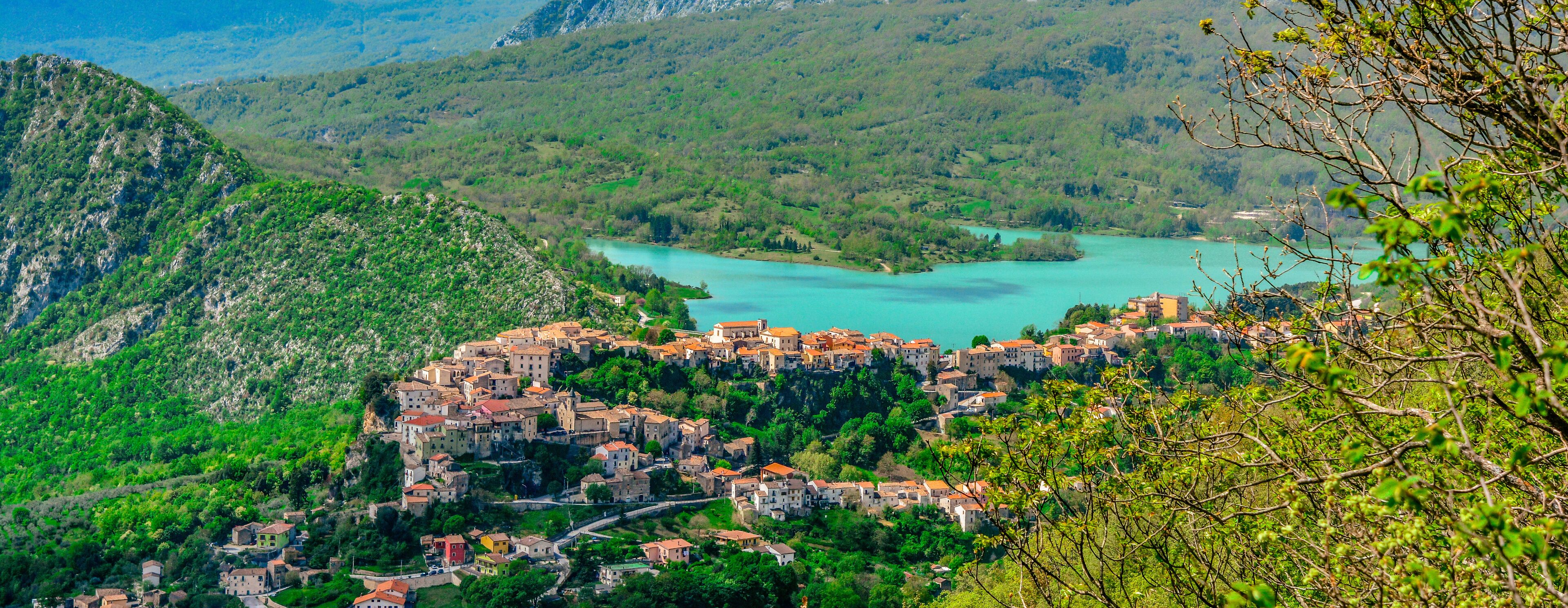 A mountainous village with terracotta-roofed buildings nestled among lush green hills and a turquoise lake.