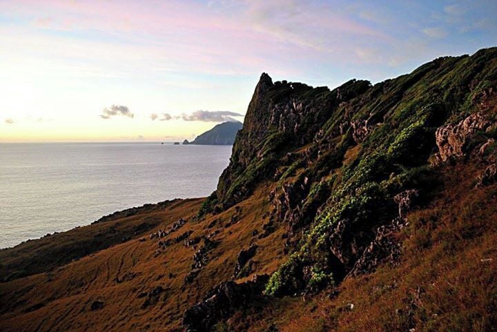 A coastal landscape with a steep cliff, grassy terrain, and a view of the sea and distant landforms.