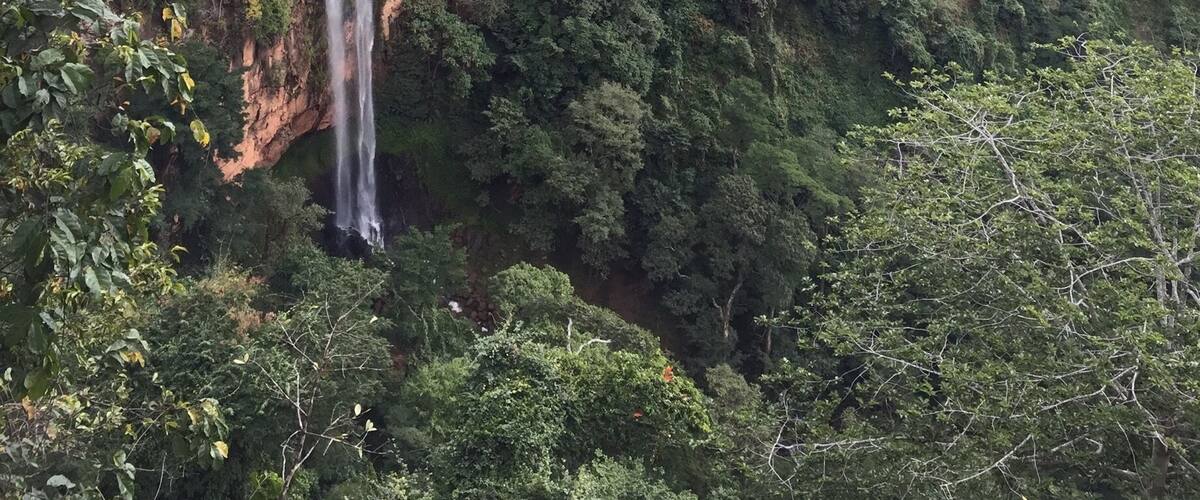 A waterfall cascading down a lush, green hillside surrounded by dense forest and distant hills.