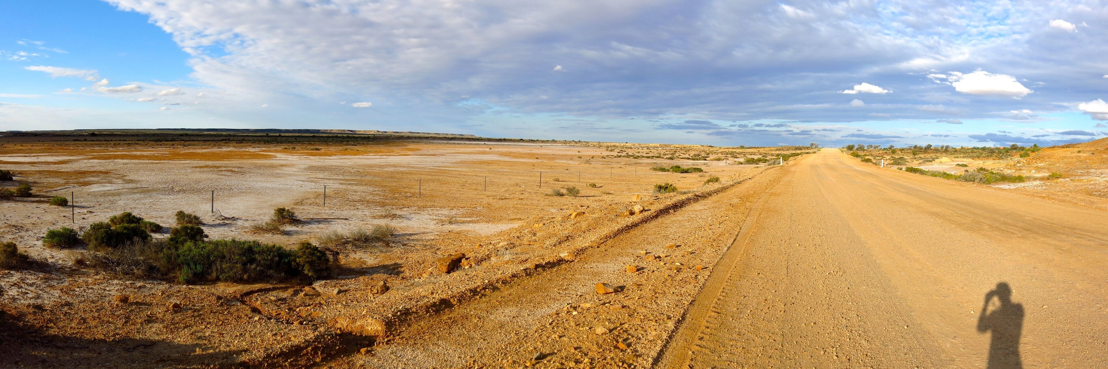 A long, straight dirt road in a barren landscape with sparse vegetation and distant hills.