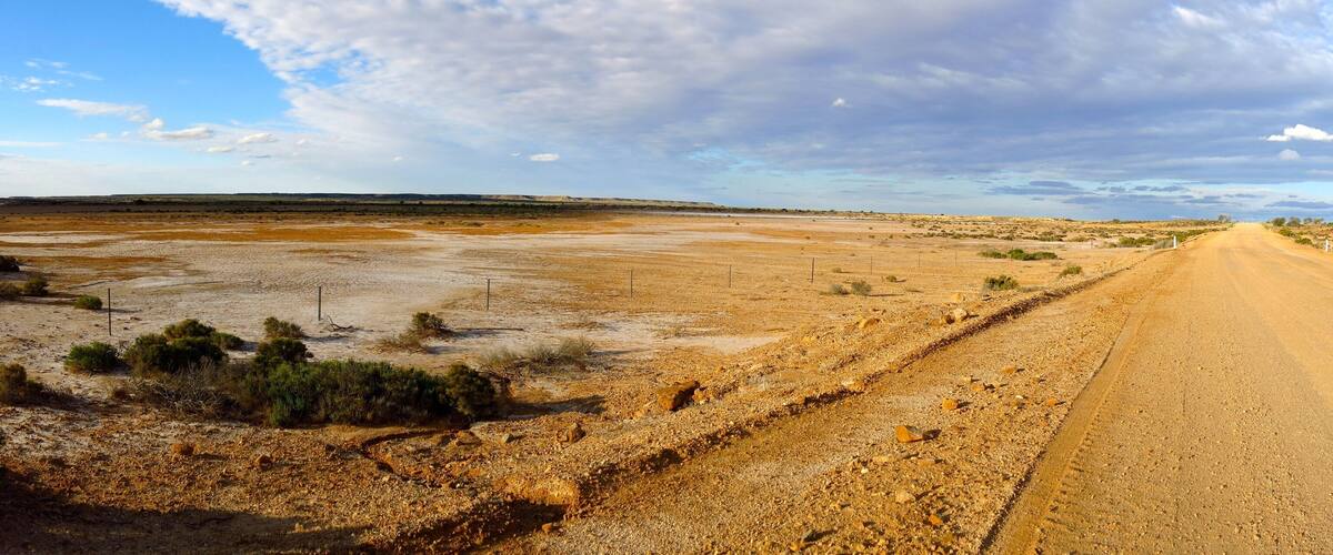 A long, straight dirt road in a barren landscape with sparse vegetation and distant hills.