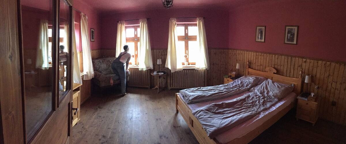 A wooden bed, a chair, a table, and a window with curtains in a room with wooden walls.