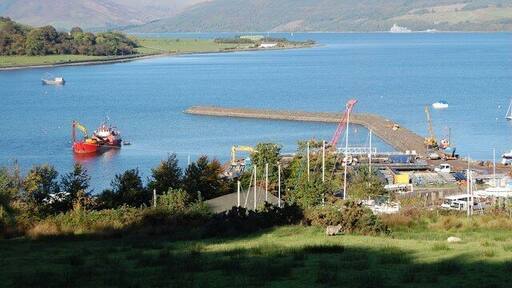 The bay at Port Bannatyne. A dredger works inside the new marina seawall. A new marina is being built at Port Bannatyne. A stone pier has been built far out into deep water, and before the pontoons can be laid, the inner water is being dredged from a ship.