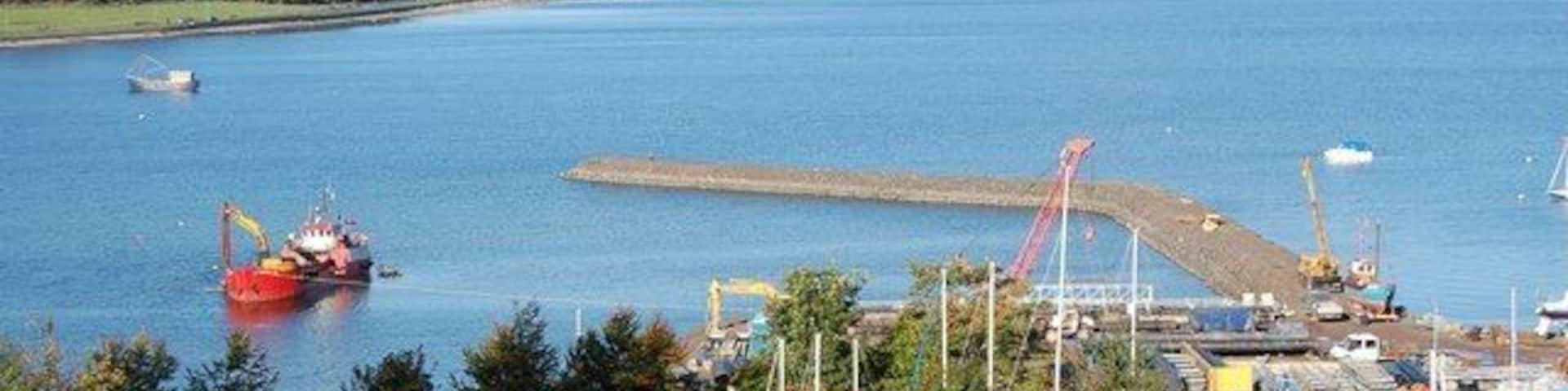 The bay at Port Bannatyne. A dredger works inside the new marina seawall. A new marina is being built at Port Bannatyne. A stone pier has been built far out into deep water, and before the pontoons can be laid, the inner water is being dredged from a ship.