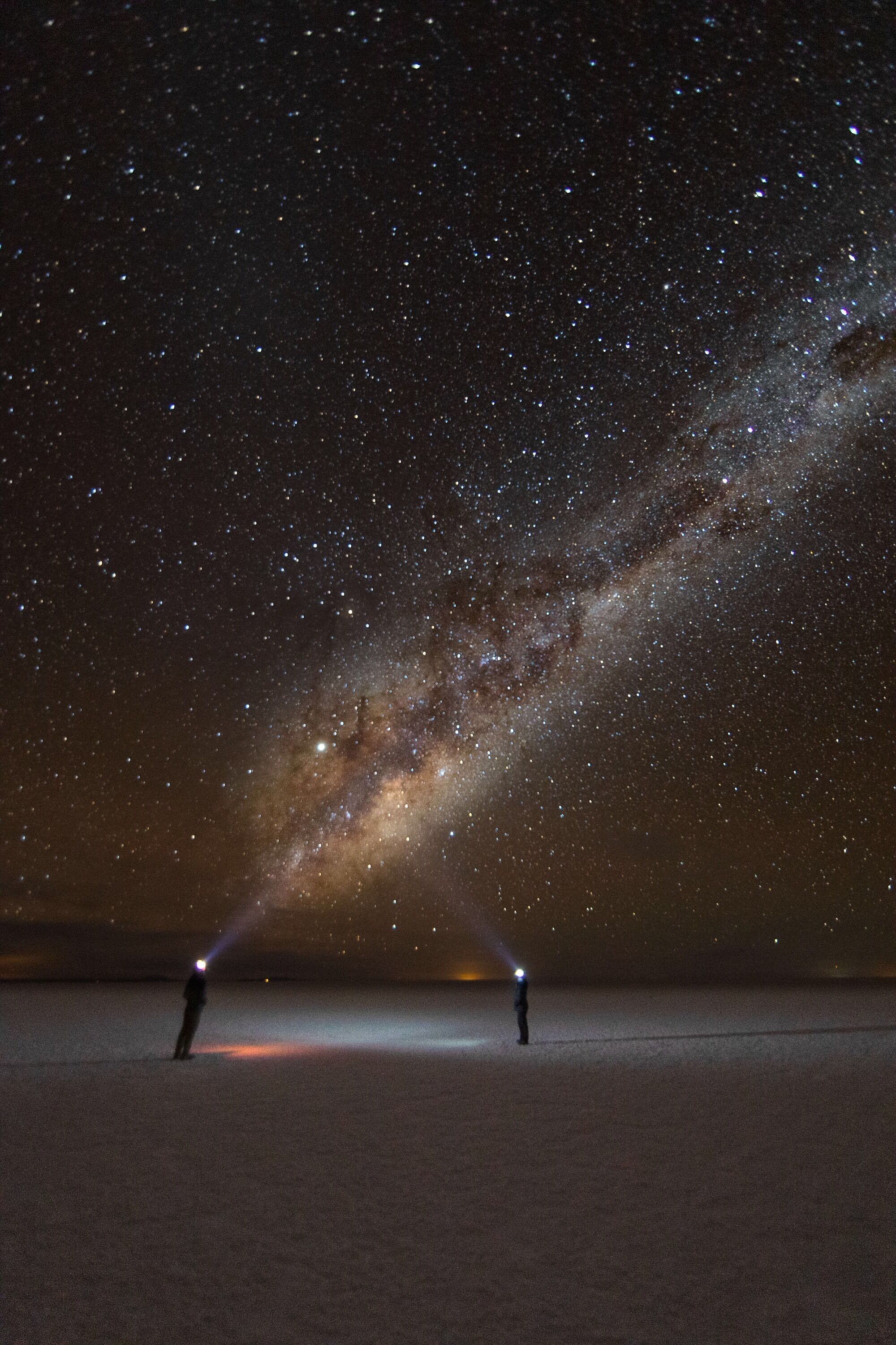 A starry night sky with a visible Milky Way and two people standing on a flat landscape.