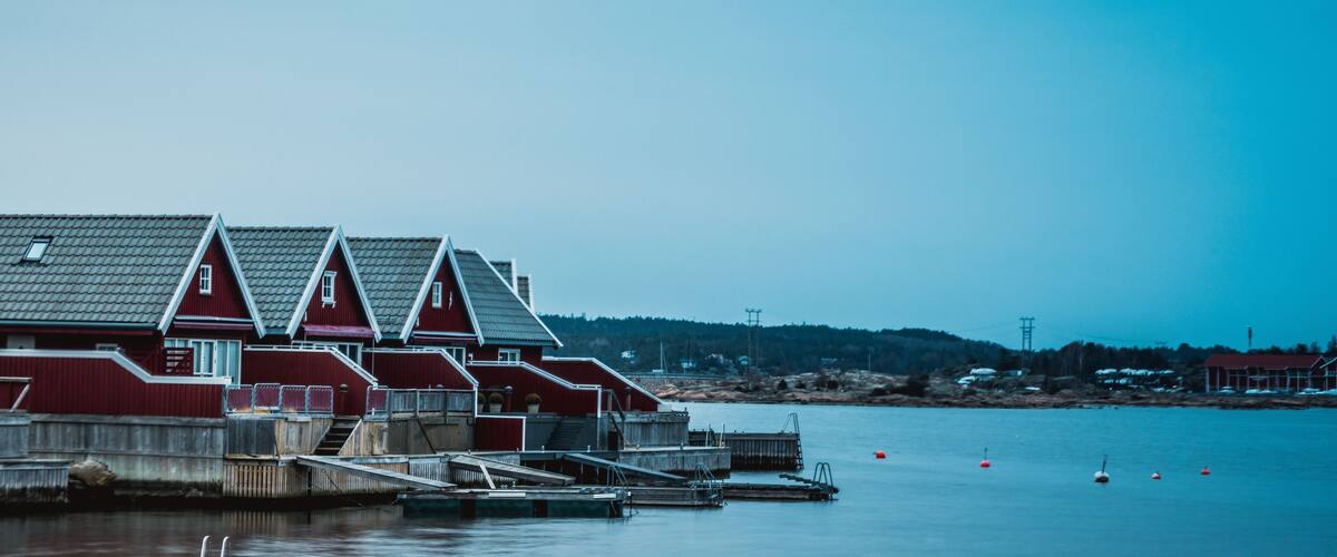 A row of red wooden houses with steep roofs by a calm body of water.