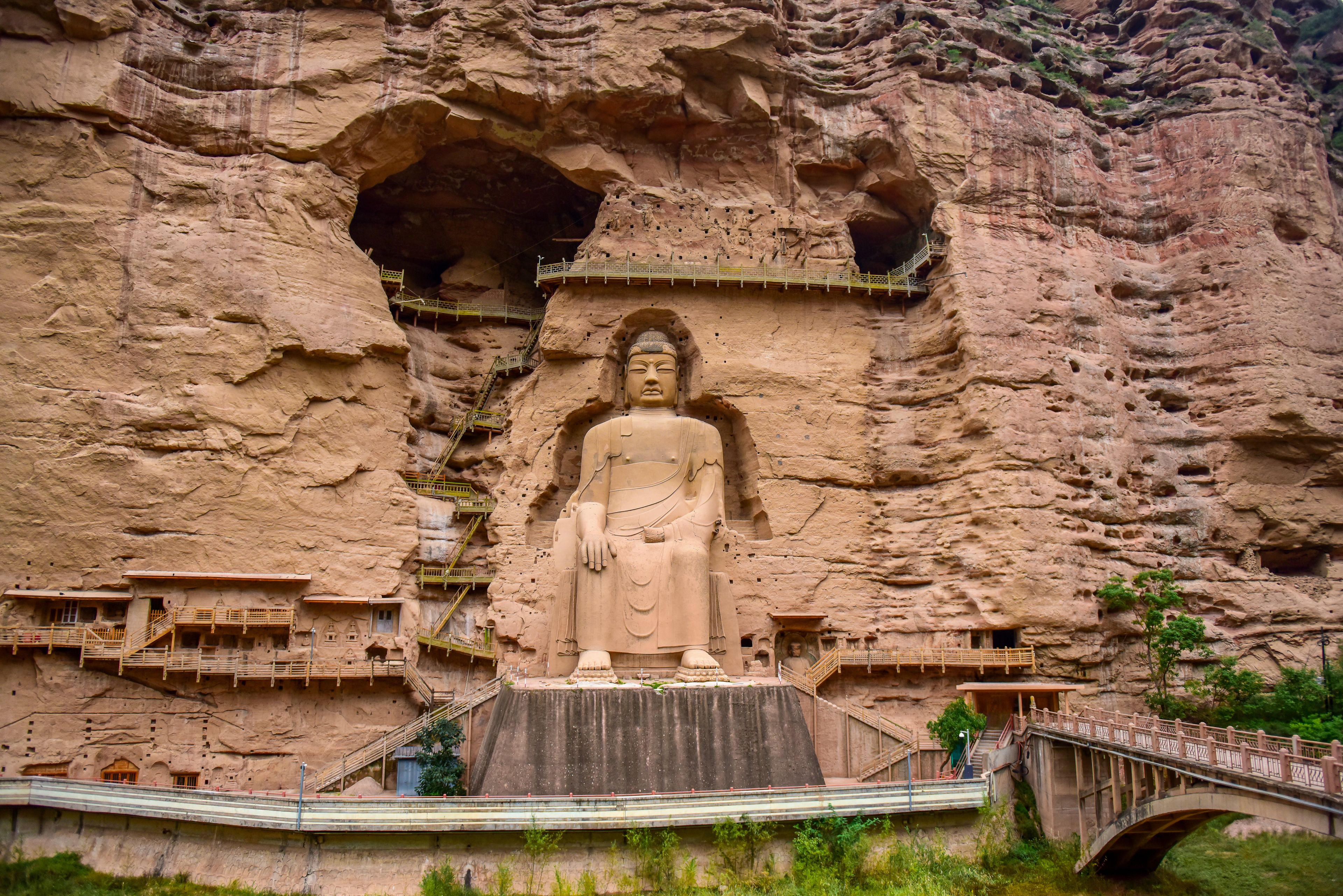 Una gran estatua de Buda tallada en la ladera de un acantilado, con un puente y senderos que conducen a ella.
