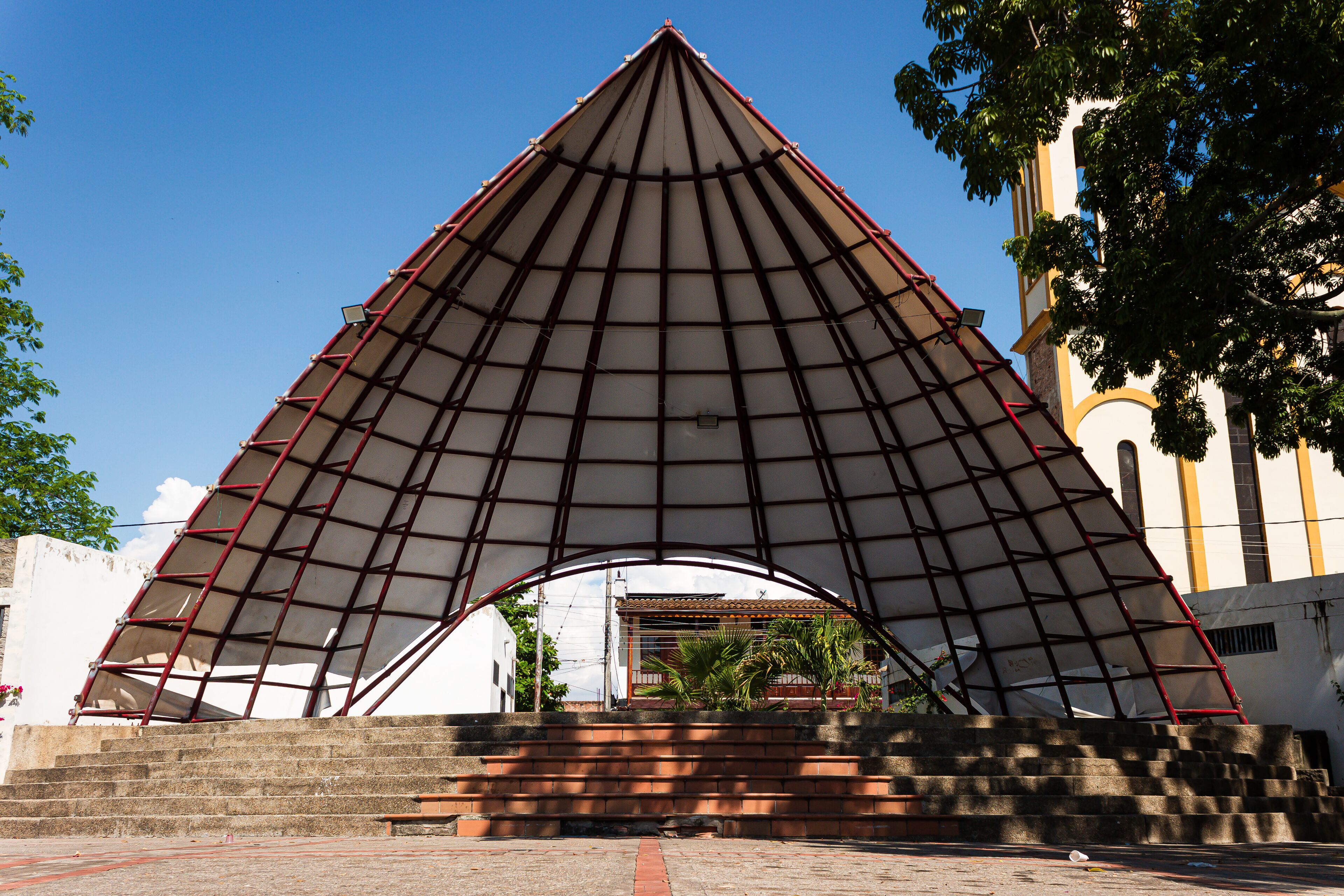 A large, triangular metal structure with a red frame and a white canopy, situated in front of a building with a clock tower.