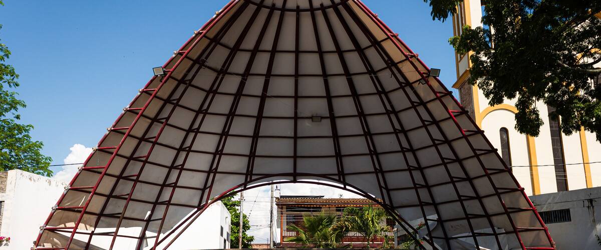 A large, triangular metal structure with a red frame and a white canopy, situated in front of a building with a clock tower.