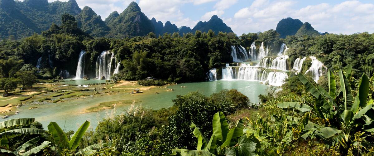 A series of waterfalls cascading into a river surrounded by lush greenery and mountains.