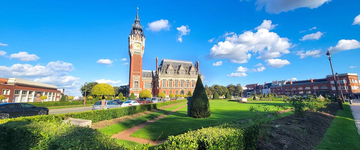 A historic building with a prominent clock tower, surrounded by well-maintained gardens and modern buildings.