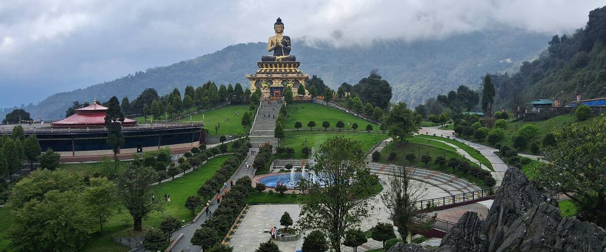 A large Buddha statue on a hilltop surrounded by lush greenery and a park with a fountain.