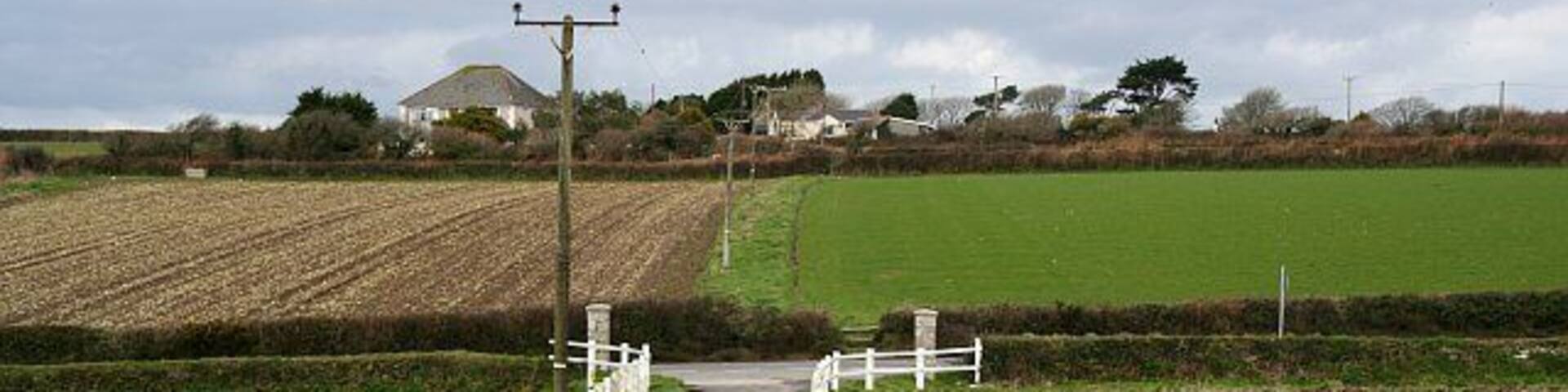 Footpath across the main road. The footpath follows the farm entrance road then crosses the A394 and onto rough grass between the fields. Almost all the signposts to this footpath have been turned around to point in a misleading direction. This is quite a common act of vandalism in rural areas.