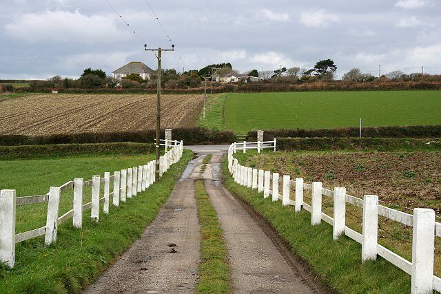 A rural road flanked by white fences, leading to a field with a farmhouse in the distance.