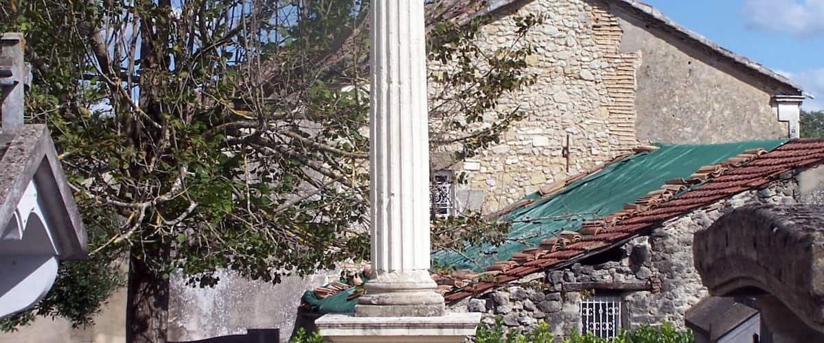 A white monument with a cross on top, surrounded by greenery and a stone building in the background.