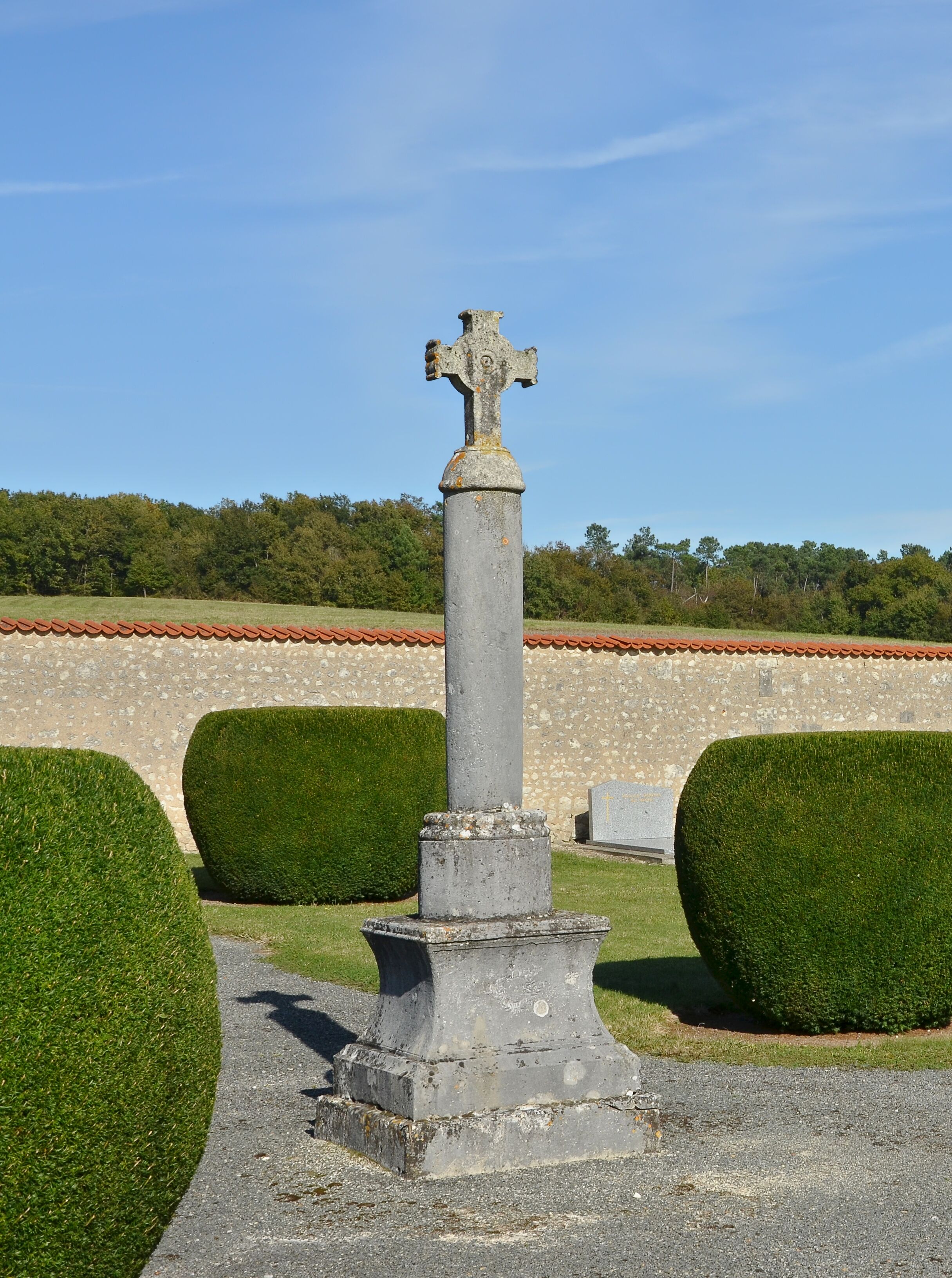 A stone cross in a cemetery with neatly trimmed hedges and a clear blue sky.