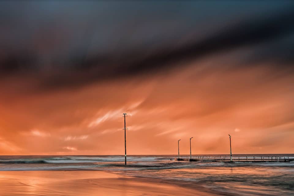 A beach with a pier extending into the ocean, under a dramatic orange sky.