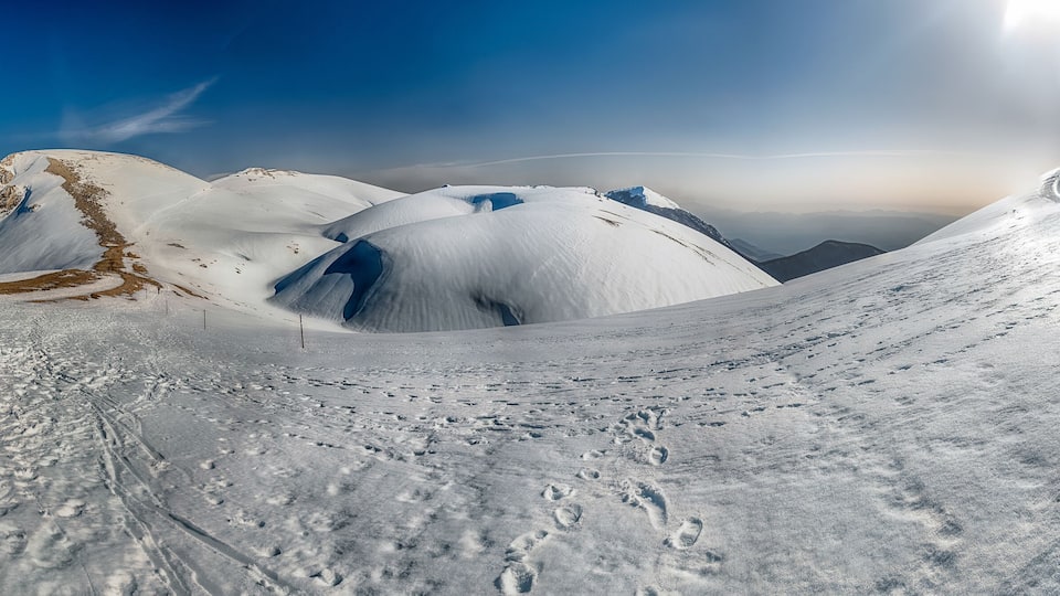 A snow-covered mountain landscape with visible footprints and a clear sky.