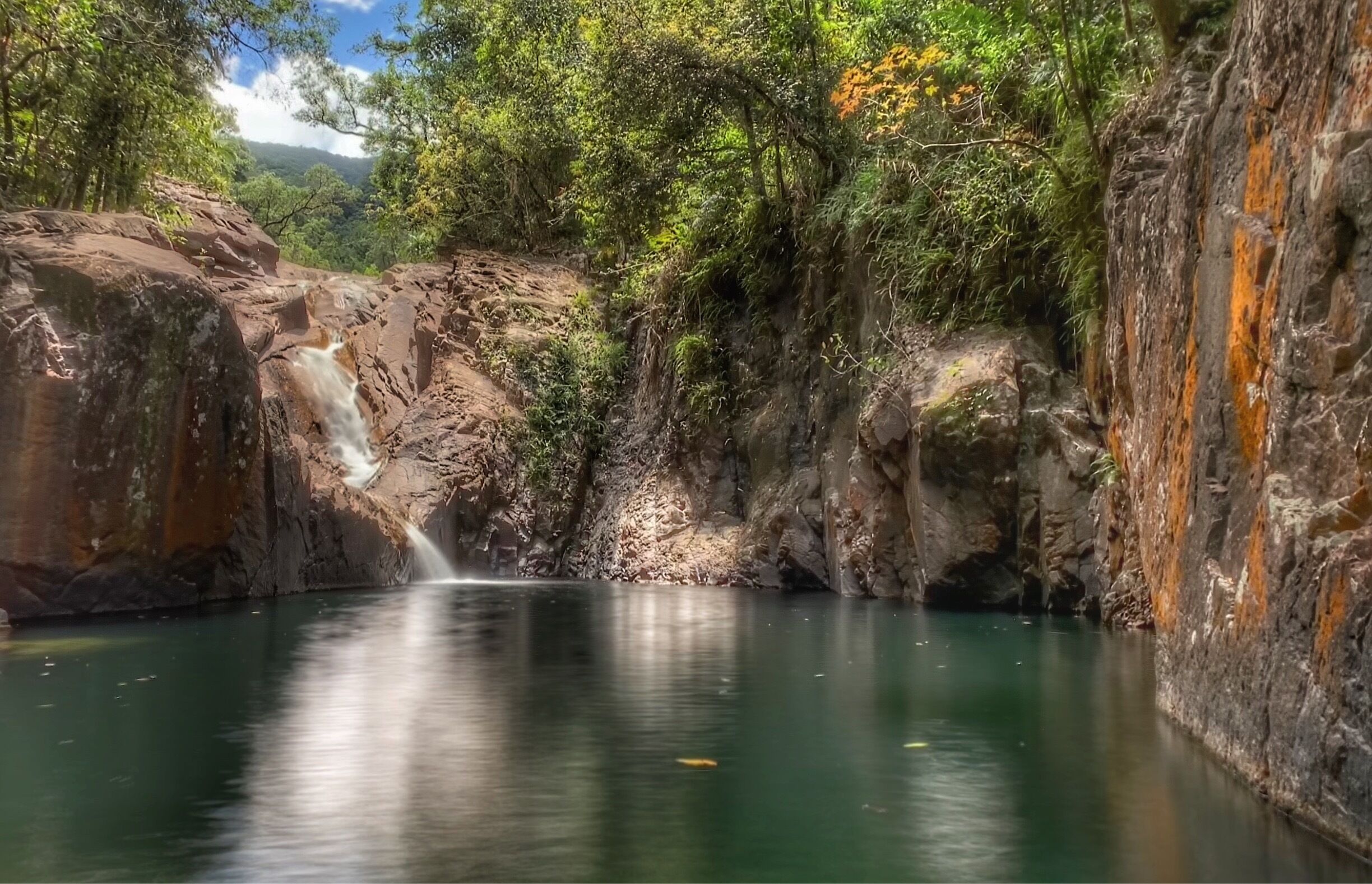 A waterfall cascading into a calm pool surrounded by rocky cliffs and dense greenery.