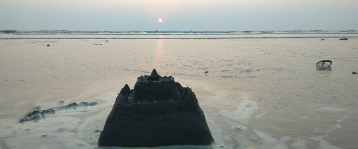 A sandy beach with a large rock formation in the foreground and the sun setting in the distance.