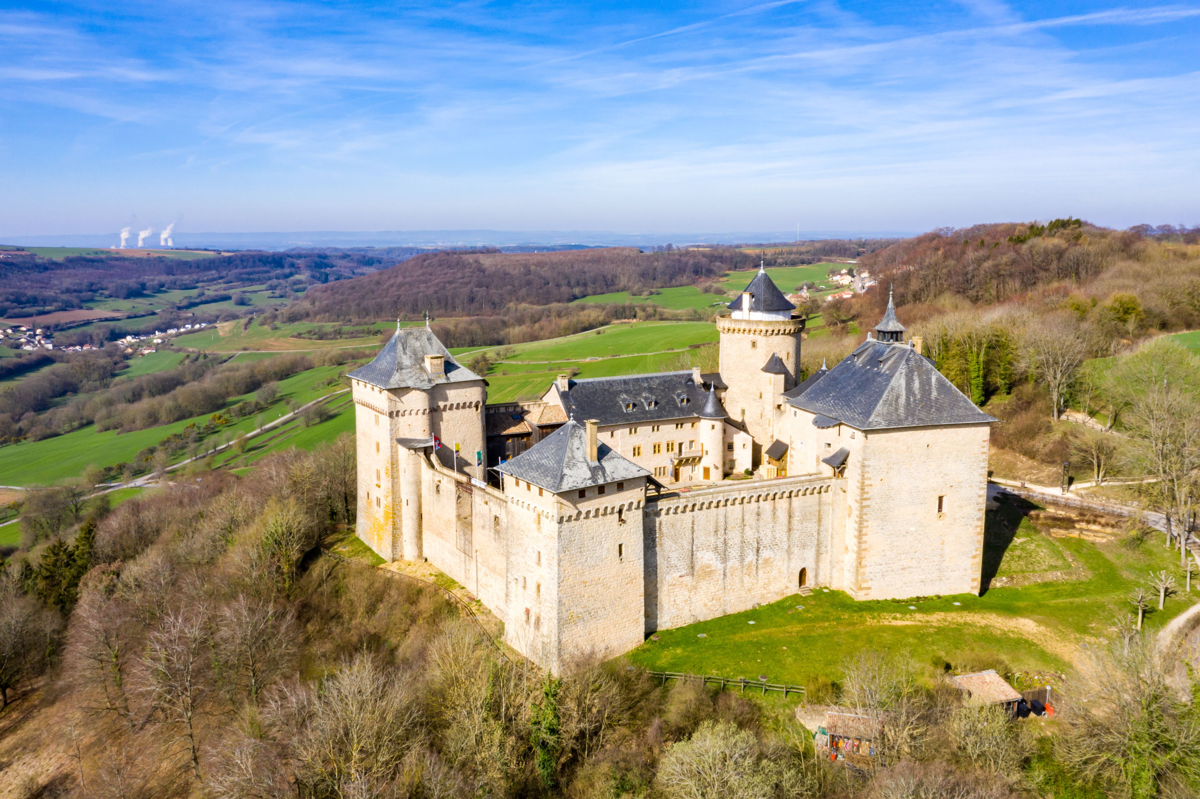 Un château doté de nombreuses tours et entouré de douves, situé dans un paysage vallonné, avec des champs verdoyants et des éoliennes visibles au loin.