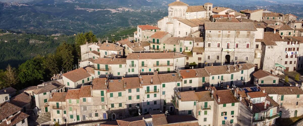 A historic town with terracotta-roofed buildings, a prominent church with a bell tower, and a mountainous backdrop.