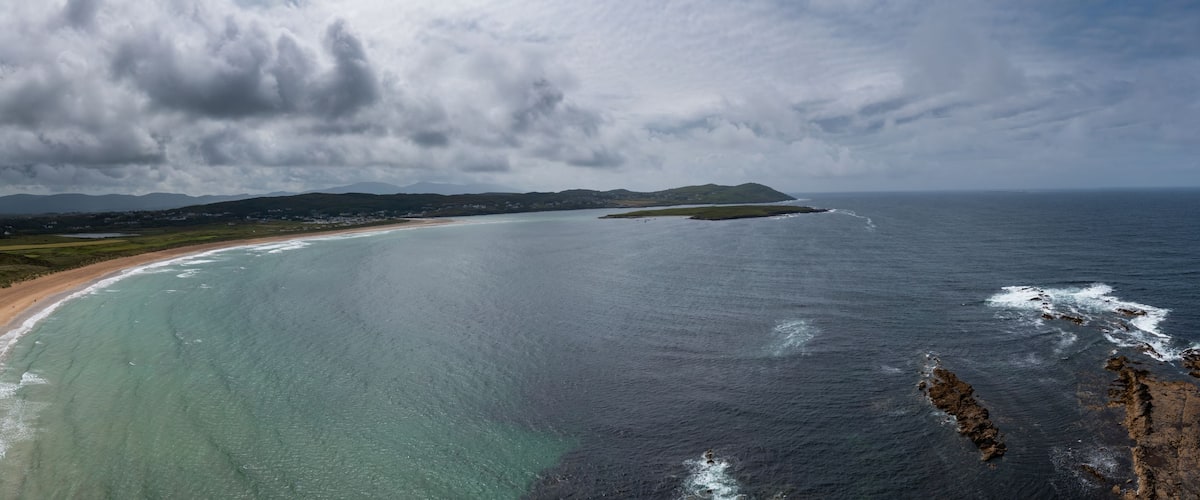 A coastal landscape with a sandy beach, calm sea, and distant mountains under a cloudy sky.