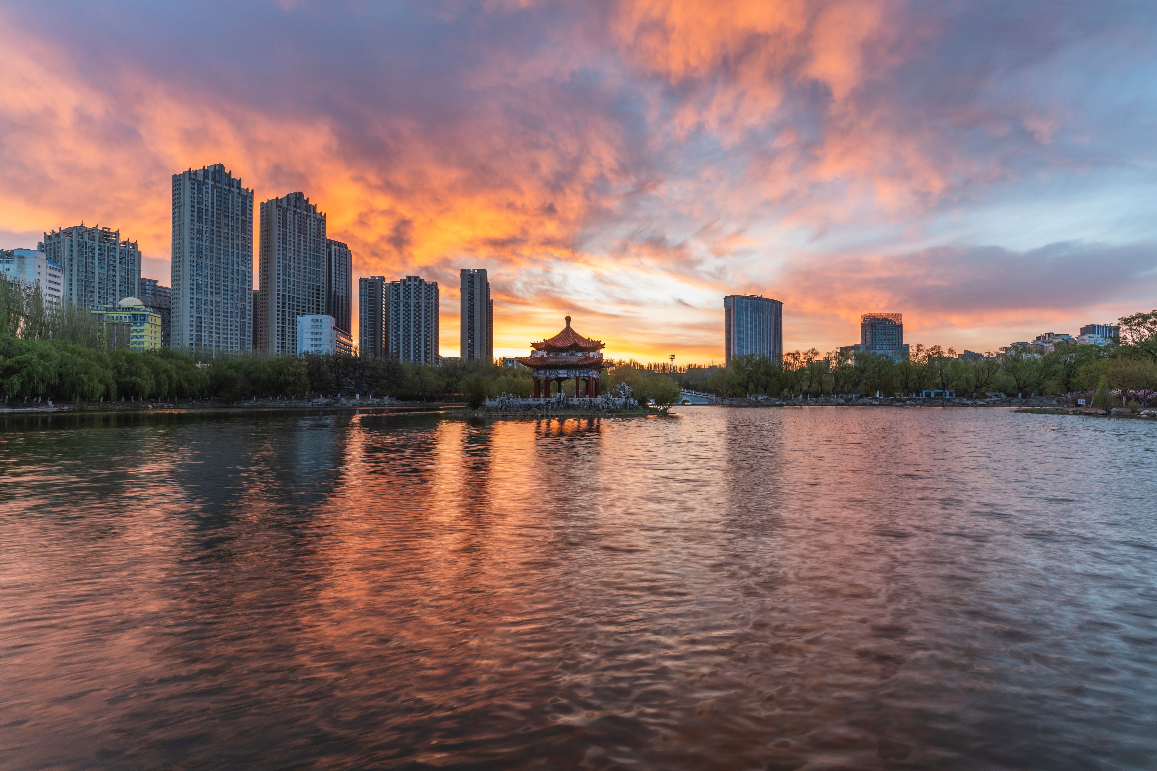 Pôr do sol sobre o horizonte urbano, com um pavilhão tradicional à beira de um rio calmo.