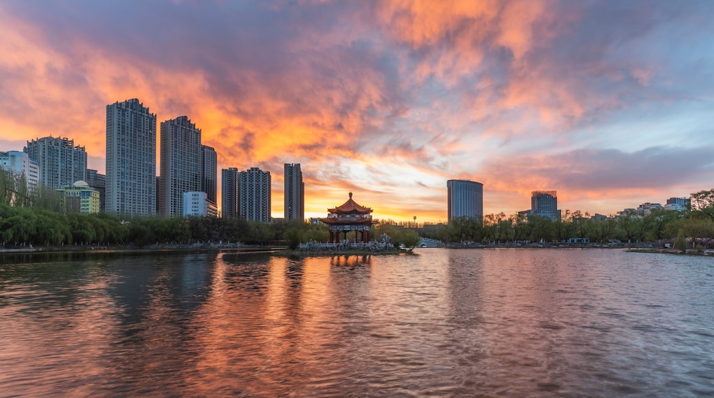 Pôr do sol sobre o horizonte urbano, com um pavilhão tradicional à beira de um rio calmo.