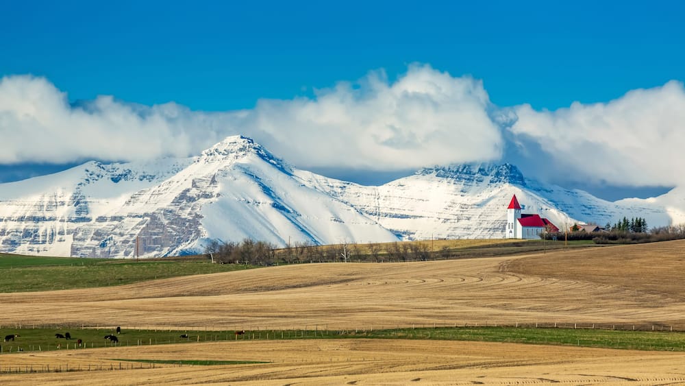 Snow-covered mountain range and brown stubble fields with white church perched on top of a hillside with blue sky and storm clouds, North of Twin Butte; Alberta, Canada