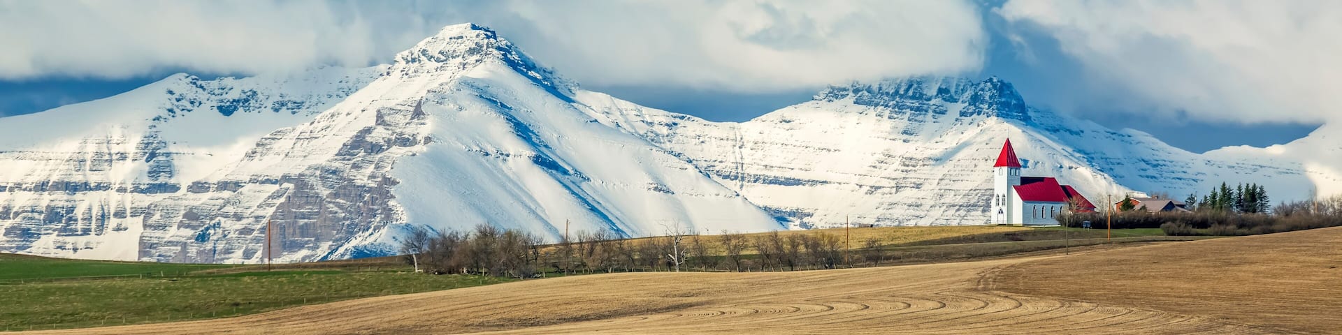 Snow-covered mountain range and brown stubble fields with white church perched on top of a hillside with blue sky and storm clouds, North of Twin Butte; Alberta, Canada