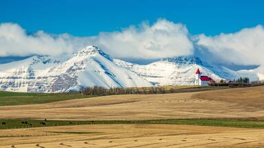 Snow-covered mountain range and brown stubble fields with white church perched on top of a hillside with blue sky and storm clouds, North of Twin Butte; Alberta, Canada