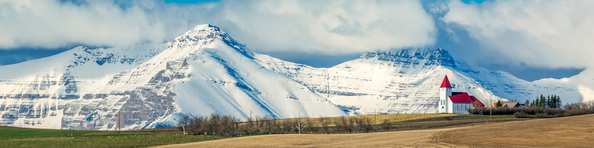 Snow-covered mountain range and brown stubble fields with white church perched on top of a hillside with blue sky and storm clouds, North of Twin Butte; Alberta, Canada