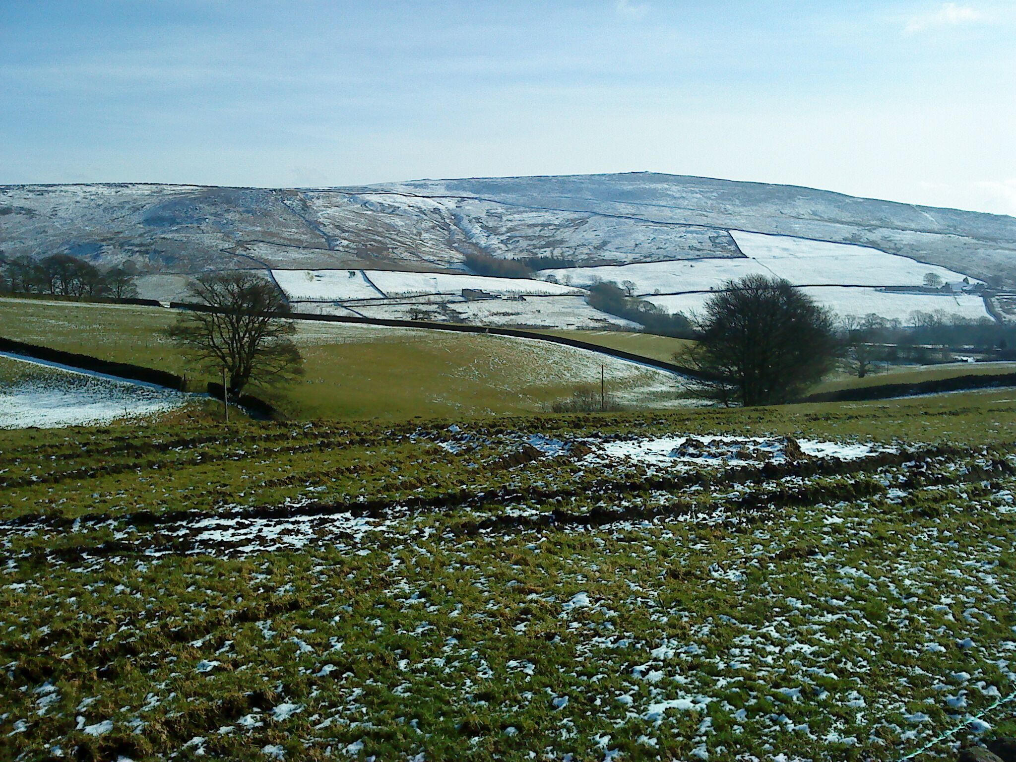 A rural landscape with snow-covered fields, a stone wall, and a hill in the background.