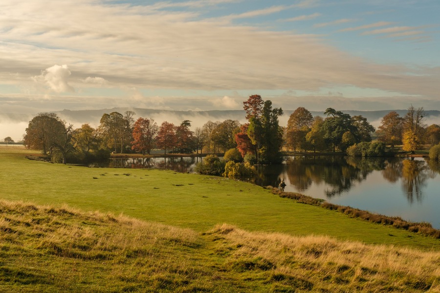 A serene lake surrounded by trees with autumn foliage, a grassy field, and a misty mountain range in the background.