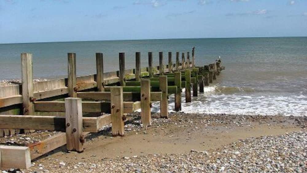 A pebbled beach with a wooden jetty extending into the sea under a partly cloudy sky.