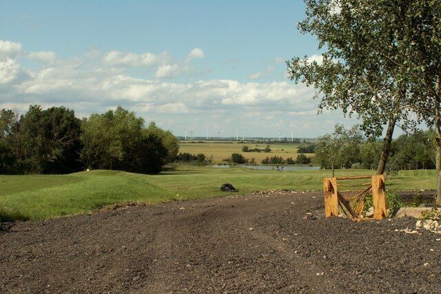 A rural landscape with a dirt path, grassy fields, a pond, and a distant wind farm.