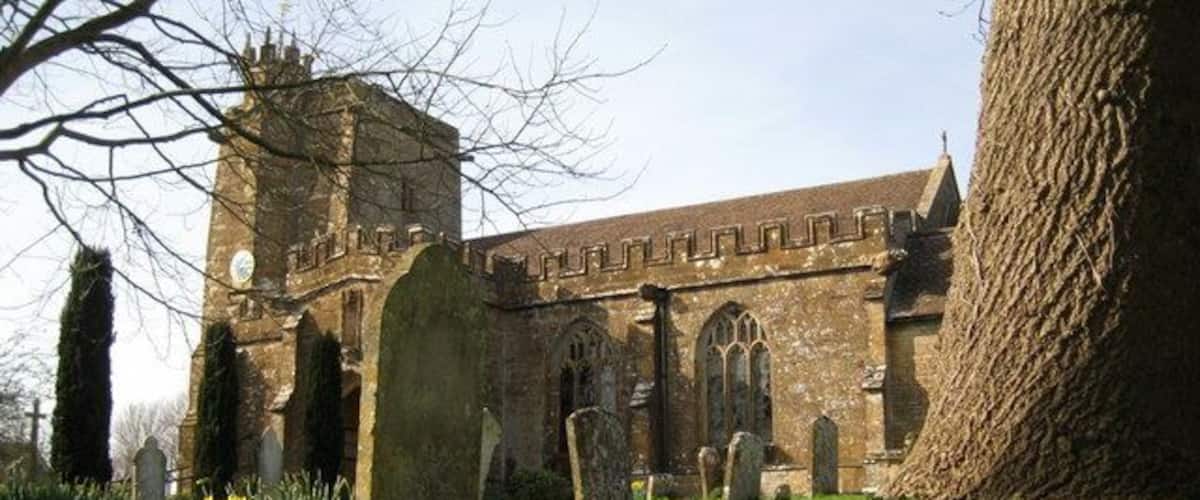 A historic church with a tall tower and arched windows, surrounded by a cemetery with headstones and blooming flowers.