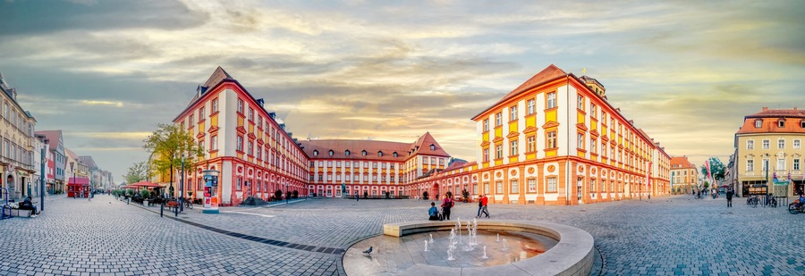 A historic square with cobblestone pavement, a central fountain, and colorful buildings with traditional architecture.