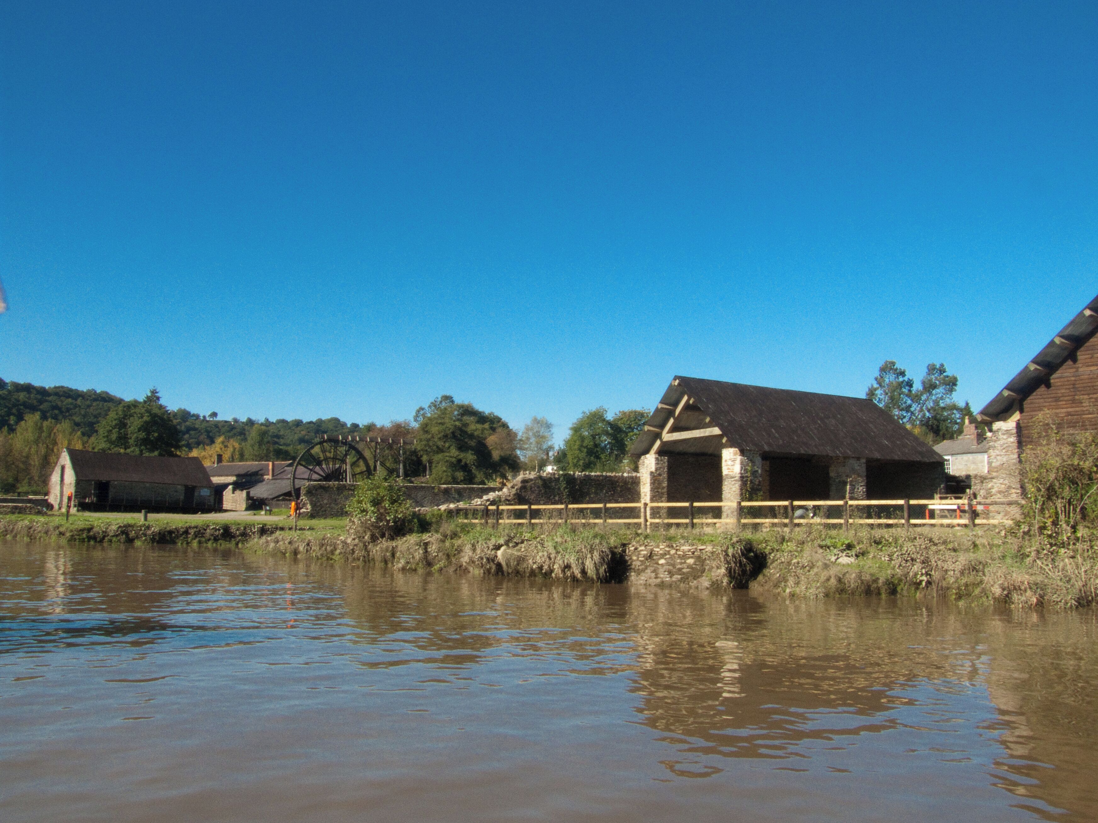 A rural landscape with a water wheel, stone buildings, and a river.