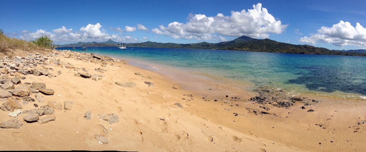A beach with clear turquoise water, rocky shoreline, and distant mountains under a blue sky with clouds.