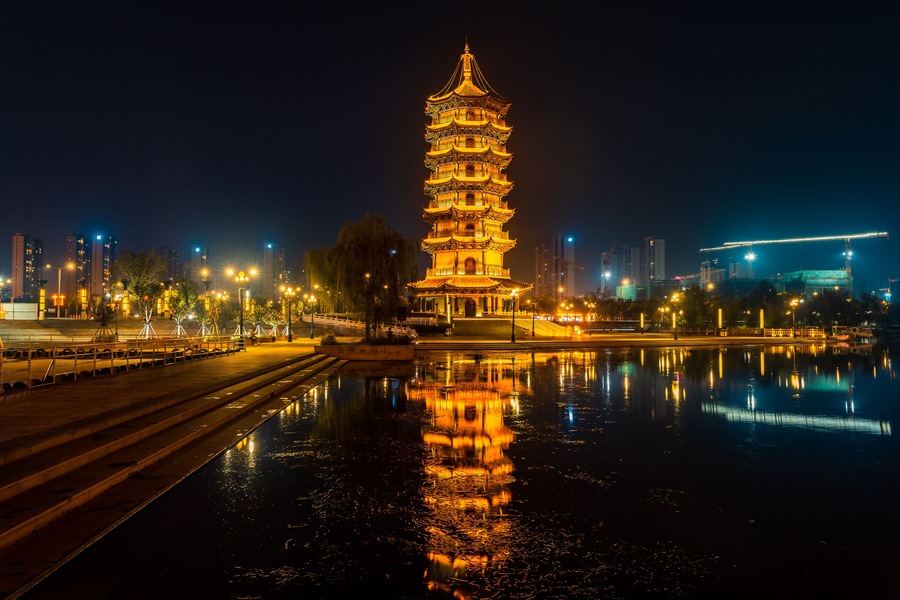 Una pagoda tradicional con varios niveles, iluminada durante la noche, reflejada en aguas tranquilas, rodeada de edificios modernos y farolas.