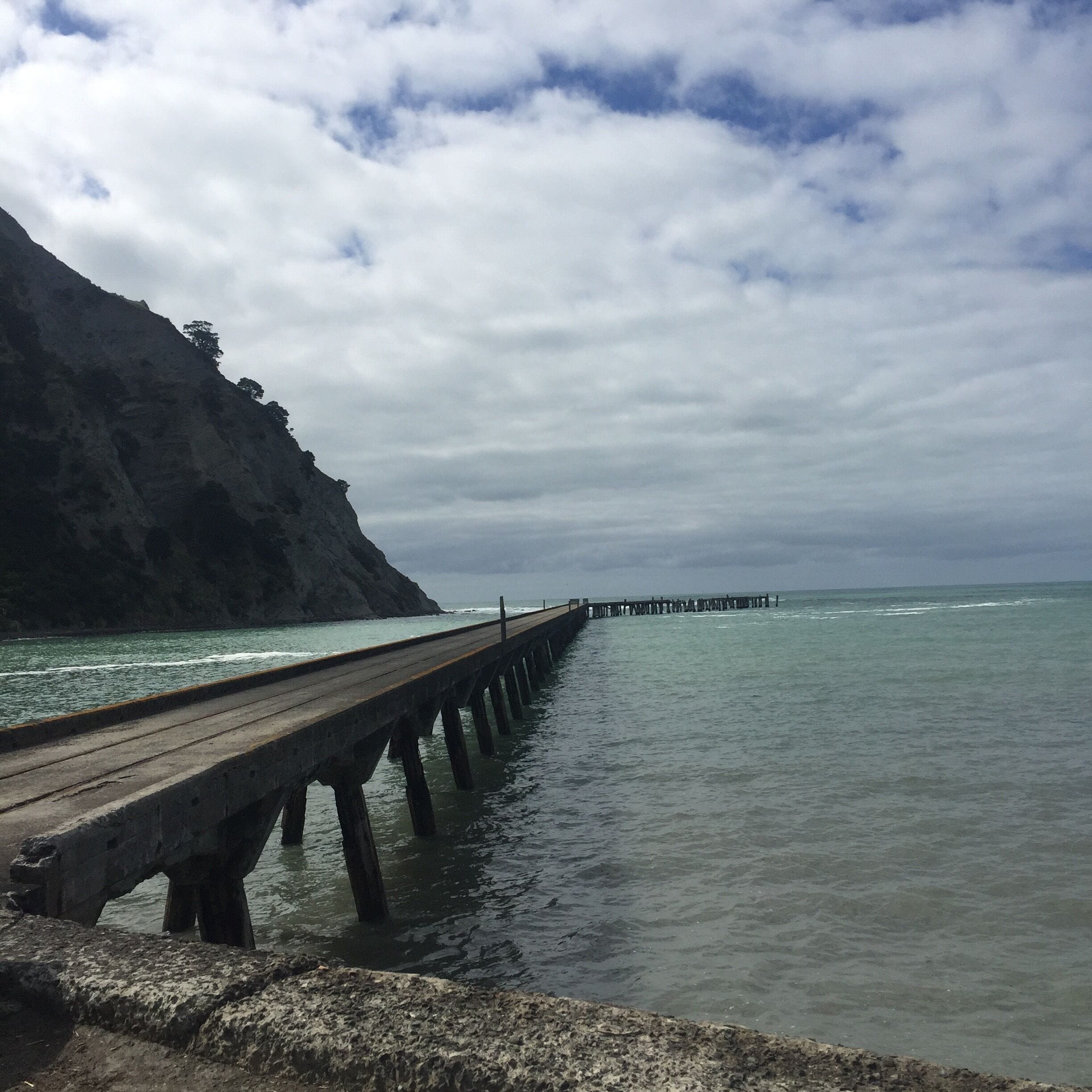 A wooden pier extending into calm waters, surrounded by rocky cliffs and a cloudy sky.