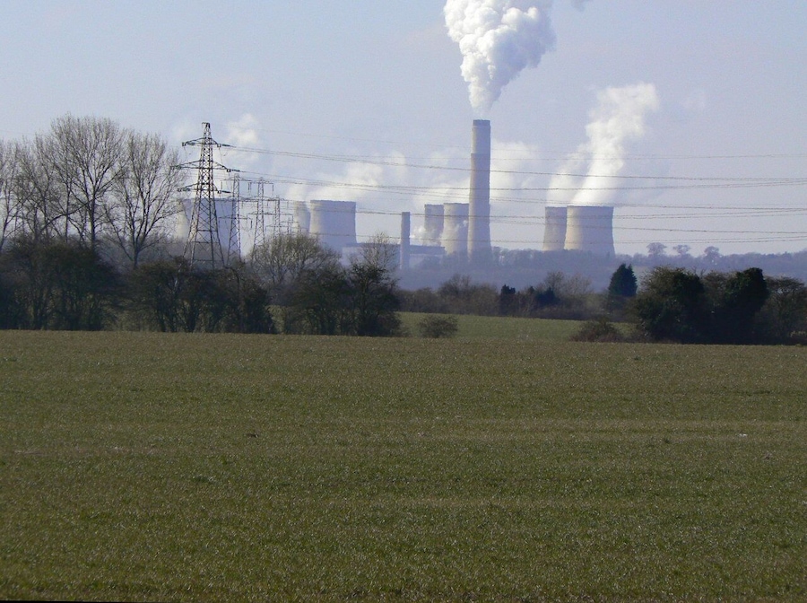 Un campo cubierto de hierba con líneas eléctricas y una fábrica distante con chimeneas altas que emiten humo.
