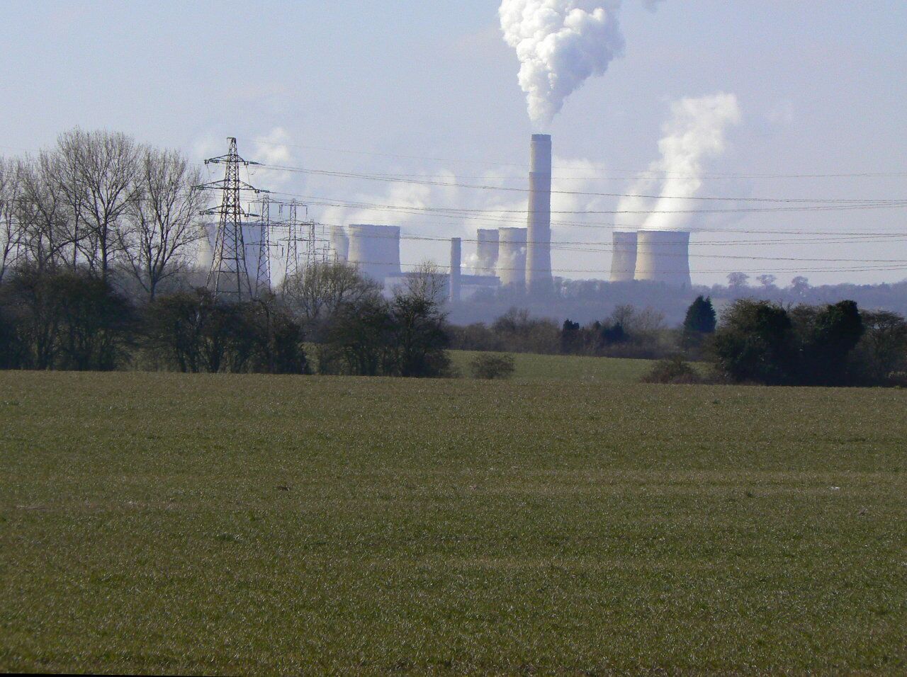 A grassy field with power lines and a distant industrial facility with tall chimneys emitting smoke.