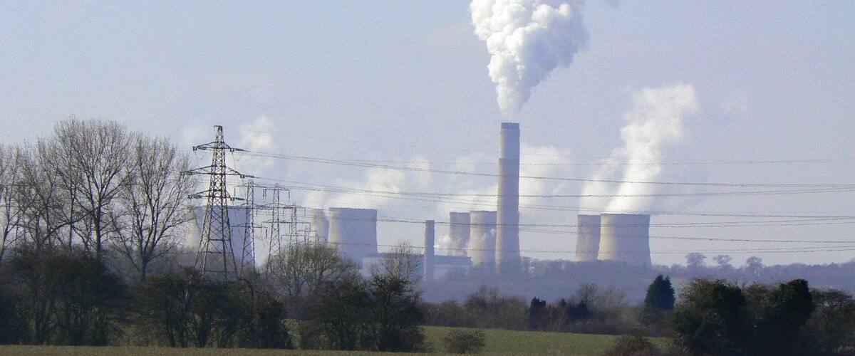 A grassy field with power lines and a distant industrial facility with tall chimneys emitting smoke.