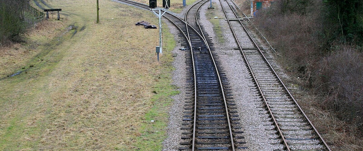 Railway tracks curving through a rural landscape with grassy fields and sparse trees.