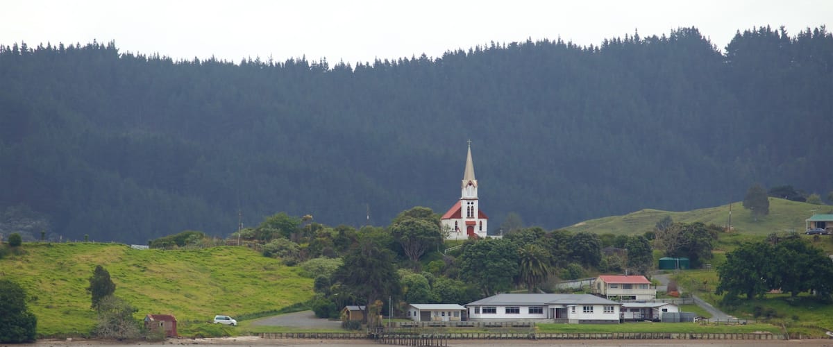 A church with a tall steeple on a grassy hill, surrounded by trees and a body of water.